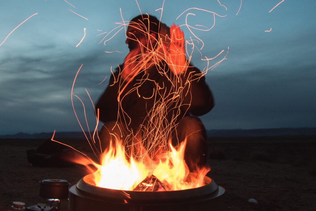 En ensam man sitter bakom en liten brasa, han håller upp sina händer framför elden och tittar åt vänster i bilden. Det är mörkt omkring. 