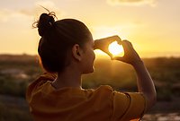 En kvinna står med ryggen mot oss framför ett öppet landskap. Det är solnedgång och hon formar ett hjärta med händerna runt solen.