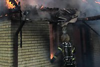 En brandman står och tittar in vid ett fönster i ett tegelhus i gult. Det brinner i rummet och vid takfoten - takplåt hänger deformerat över brandmannen...